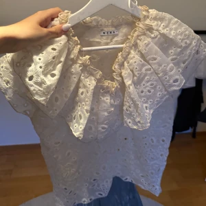 Broderad blus med volang från Wera - Superfin ljusbeige blus från Wera med broderade blommor och hålmönster. Stora volanger runt halsen ger en romantisk vibe. Perfekt till jeans eller kjol för en söt look. Luftig och lätt, passar grymt till varmare dagar.