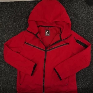 Röd Nike vindjacka med luva Medium size - Nike vindjacka i rött med svarta detaljer och hel dragkedja. Jackan har huva, lång ärm och en snygg svart rand över bröstet samt en praktisk ficka med dragkedja på ärmen. Perfekt för dig som gillar sportig och clean stil.