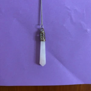 Halsband med rosa kristallhänge - Snyggt halsband med en lång, spetsig rosa kristall som jag har för mig är bergskristall som hänge. Kristallen sitter i ett detaljerat metallfäste med antik känsla och hänger i en tunn silverfärgad kedja. Perfekt för dig som gillar unika smycken med boho-vibe.