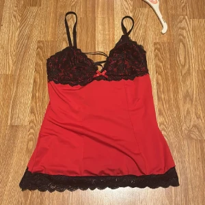 Röd och svart spetslinnetopp med snörning - Säljer en röd linnetopp med svarta spetsdetaljer och smala axelband. Toppen har dekorativ snörning i ryggen och en liten rosett framtill. Spetsen finns både vid bysten och längs nederkanten, vilket ger en extra snygg touch. Perfekt för dig som gillar en lite mer vågad stil. Tryck ej köp nu