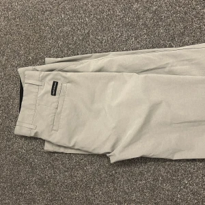 Golf byxor från peak performance  - Snygga beige golfbyxor från peak med klassisk rak passform. Byxorna har bälteshällor, knapp och dragkedja fram samt bakficka med diskret logotyp. Perfekta för en clean och stilren look. Storlek 29/32