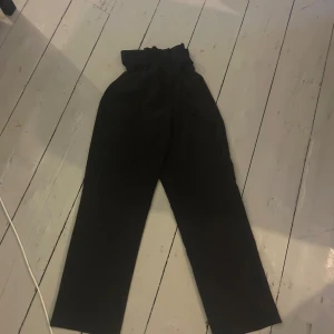 Svarta kostymbyxor med bälte - Snygga svarta kostymbyxor med hög midja och papperspåse-linning. Byxorna har ett matchande tygbälte och stängs med dragkedja och hyska. Raka ben och stilren look, perfekta till en trendig outfit.