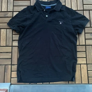 Svart pikétröja från GANT, stl S - Klassisk svart pikétröja från GANT i storlek S. Tröjan har korta ärmar, krage med knappar och broderad GANT-logga på bröstet. Perfekt för en clean och stilren look. Lite solblekt och ett hål vid knapparna. 