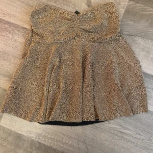 Glittrig guld tubtopp från Missay - Supersnygg glittrig tubtopp i guld från Missay. Toppen har bandeau-modell med lätt rynkad byst och ett flowigt fall nedtill. Perfekt för dig som vill sticka ut med metallic-vibes och cool textur. Säljs med prislapp kvar.