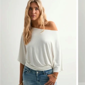 Vit offshoulder topp från Nelly - Säljer en vit offshoulder topp från Nelly med lös passform och trekvartsärm. Toppen har en snygg, avslappnad siluett och är tillverkad i mjukt material som faller fint över axeln. Perfekt för dig som gillar en stilren och trendig look.