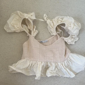 Beige volangtopp från Zara - Supersöt beige topp från Zara med smockad överdel och volang nertill. Axelbanden är breda och har volanger samt knyt, vilket ger en romantisk vibe. Perfekt för varma dagar och riktigt snygg till jeans eller kjol.