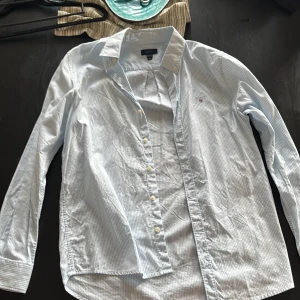 Blåvit randig skjorta från GANT - Snygg blåvit randig skjorta från GANT med klassisk krage och knappar framtill. Skjortan har lång ärm och broderad GANT-logga på bröstet. Perfekt för dig som gillar stilrena och tidlösa plagg. Tillverkad i mjuk bomull för en skön känsla.