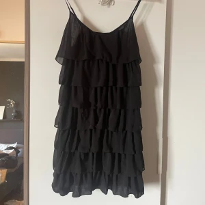 Svart volangklänning med tunna axelband - Supersöt svart kortklänning med flera lager volanger och tunna axelband. Perfekt för dig som gillar en lekfull och trendig stil. Lätt och luftig känsla, passar till många olika tillfällen.