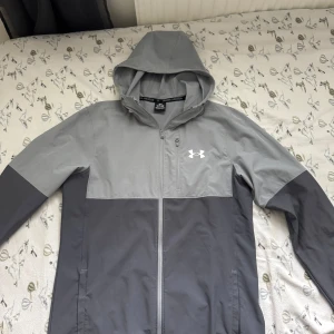 Grå vindjacka från Under Armour - Snygg tvåfärgad grå vindjacka från Under Armour med huva och hel dragkedja. Jackan har en bröstficka med dragkedja och logga på bröstet. Perfekt för blåsiga dagar och sportiga outfits. En liten defekt på höger arm som ni ser själva har jag lagt upp en bild på  det. Annars topp klass 