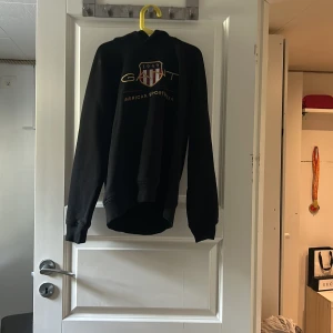 Svart hoodie från GANT med tryck (äkta) - Svart hoodie från GANT med klassiskt tryck och logga framtill i guld och rött. Tröjan har huva, ribbade muddar och är tillverkad i mjuk bomullsmix. Perfekt för en chill och sportig look.