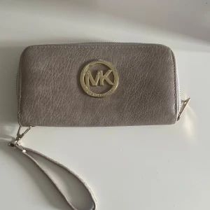 Beige plånbok Michael Kors skinn - Snygg beige plånbok från Michael Kors i skinn med guldigt MK-emblem framtill. Plånboken har dragkedja runtom, flera kortfack, myntfack med dragkedja och en praktisk handledsrem. Perfekt för att hålla koll på allt viktigt med stil.