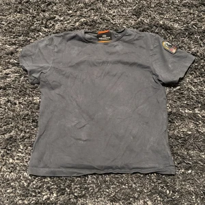 Mörkgrå t-shirt från Parajumpers - Snygg mörkgrå t-shirt från Parajumpers med klassisk passform och rund hals. På vänster ärm sitter ett broderat PJS-märke som ger en cool detalj. Tillverkad i mjuk bomull, perfekt för en avslappnad stil. Äkta!!