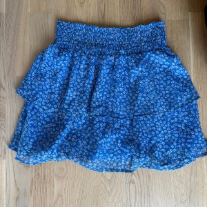 Blå blommig volangkjol med resår - Supersöt blå kortkjol med vitt blommönster och smockresår i midjan. Kjolen har volangdetaljer och är luftig, perfekt för varma dagar. Passar till vardagsanvändning och finare tillfällen. Kjolen kommer inte till användning längre. Storlek M men passar mig som bär S. 