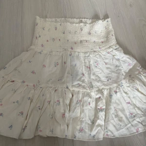 Vit blommig kjol från Lager 157 - Supersöt kort kjol i vitt med prislappen kvar från lager 157 med små blommor i rosa och blått. Kjolen har smock i midjan vilket ger en skön och stretchig passform, perfekt för sommaren!