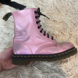 Metallic rosa Dr. Martens boots - Säljer ett par ikoniska Dr. Martens boots i metallic rosa skinn med svarta snören och klassisk gul söm runt sulan. Skorna har rund tå och platt sula, perfekt för dig som vill sticka ut med en cool färg. Grymma till jeans eller kjol! Något att notera är dock att skorna har använts väl och är lite slita, se bilder, därmed kan priset diskuteras!