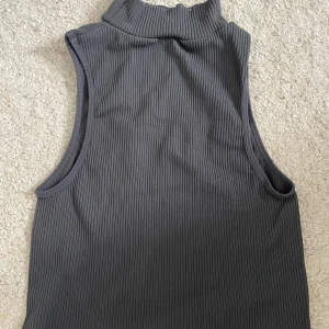 Grå ribbad polotopp ärmlös från Zara - Säljer en stilren grå ribbad polotopp utan ärmar från Zara. Toppen har hög hals och är tillverkad i ett mjukt, stretchigt material som sitter snyggt på kroppen. Perfekt att matcha med jeans eller kjol för en clean look.