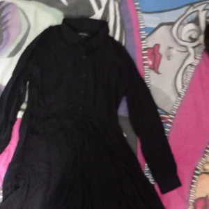 Svart långärmad klänning från Amisu - Svart långärmad klänning från Amisu i storlek 34. Klänningen har klassisk skjortkrage, knappar framtill och ett lätt utställt fall. Perfekt för dig som gillar en stilren look med enkel design.