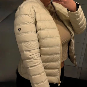 Beige pufferjacka från Rockandblue - Snygg beige pufferjacka från Rockandblue med raka quiltade sömmar och guldfärgade dragkedjor. Jackan har en stilren look utan krage, två fickor med dragkedja och Rockandblue-logga på ärmen. Perfekt för dig som vill ha en clean och trendig jacka till höst och vår.