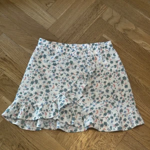 Blommig volangkjol  - Supersöt kort kjol med blommigt mönster i grönt, rosa och blått på vit botten. Kjolen har volangkant och omlott-look framtill som ger en extra gullig touch. Perfekt för varma dagar och lätt att matcha med en enkel topp.💕