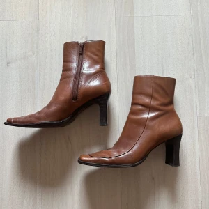 Bruna skinnstövletter från Bronx - Snygga bruna stövletter i skinn från Bronx med fyrkantig tå och markerad klack. Dragkedja på insidan för enkel påtagning och stilren design som ger en retrostil.