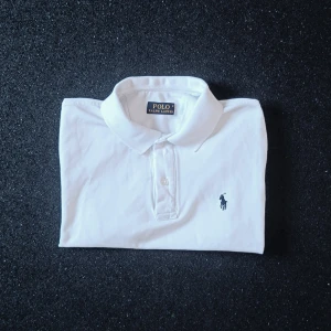Ralph Lauren Piké Vit storlek S-M - Klassisk vit pikétröja från Ralph Lauren med broderad svart logga på bröstet. Tröjan har krage, korta ärmar och knäppning framtill. Tillverkad i mjuk bomull som känns skön mot huden. Perfekt för en clean och stilren look.