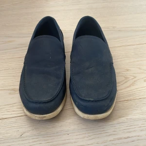 Mörkblå mockaloafers med vit sula - Snygga mörkblå loafers i mocka med stilren design och vit platt sula. Skorna har en klassisk form med rund tå och passar perfekt till både jeans och chinos för en clean lock. Endast använda två gånger. De ser lite smutsiga ut på bilderna men allting går bort vid en enkel tvätt. Inga defekter och de är i helt nyskick i övrigt från smutsen.