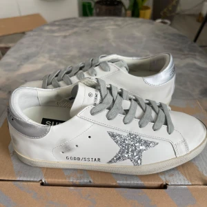 Golden Goose vita sneakers med glitterstjärna - Snygga vita sneakers från Golden Goose med silverfärgade detaljer och en glittrig stjärna på sidan. Skorna har grå snörning och en klassisk låg profil. Perfekta för dig som vill sticka ut med en cool och trendig look.