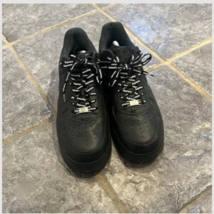 Nike Air Force 1 x Supreme svarta sneakers - Svarta Nike Air Force 1 sneakers i samarbete med Supreme. Skorna har klassisk låg siluett, svart skinn och Supreme-logga på sidan. Svarta och vita skosnören med Supreme-tryck. Perfekta för dig som gillar streetwear och vill sticka ut.