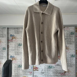 Beige stickad kofta från MNG - Snygg beige stickad kofta från MNG med klassisk krage och knappar framtill. Koftan har lång ärm och är ribbstickad för en clean och stilren look. Perfekt att slänga över en t-shirt eller linne när det blir lite kyligt.