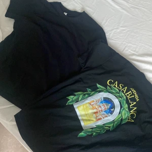 Svart Avenida Casablanca t-shirt - Svart t-shirt med färgglatt tryck på ryggen som föreställer en byggnad och gröna blad, samt texten 'Avenida Casablanca' i gult. Klassisk passform och rund hals. Perfekt för dig som vill sticka ut med en unik och konstnärlig vibe. Finns två stycken styckpris helt nya