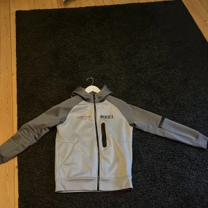 Grå zip hoodie från JD sports Hoodrich med luva - Snygg grå zip hoodie från Hoodrich med svart tryck och röda detaljer. Jackan har huva, hel dragkedja framtill och en cool bröstficka med svart dragkedja. Mjuk insida och sportig look, perfekt för dig som gillar streetwear.