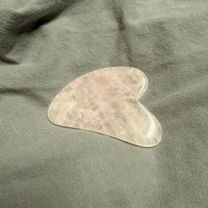 Rosa gua sha-sten i rosékvarts - Säljer en gua sha-sten i äkta rosékvarts med mjukt rundad form och hjärtliknande design. Perfekt för ansiktsmassage och att boosta hudvårdsrutinen. Stenen är ljusrosa med naturliga marmoreringar och känns sval och lyxig mot huden. Den är oanvänd 