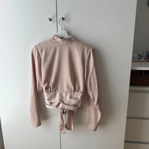 Champagnefärgad omlottopp Gina Tricot - Supersnygg champagnefärgad omlottopp från Gina Tricot i satinliknande material. Toppen har lång ärm, hög krage med knappar och knyt i midjan som ger en elegant look. Perfekt för dig som vill sticka ut med en stilren och trendig topp.