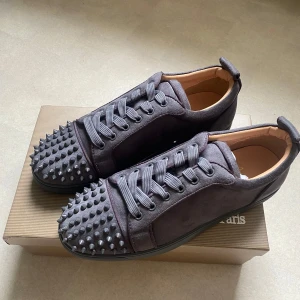 Grå mocka sneakers från Louboutin - Snygga grå sneakers i mocka från Louboutin med ikoniska röda sulor och coola nitar på tån. Skorna har snörning och en platt sula. Perfekta för dig som vill sticka ut med en edgy och lyxig stil.