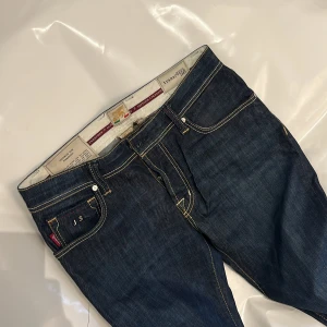 Mörkblå jeansbyxor med detaljer - Snygga mörkblå jeansbyxor från Tramarasso med kontrastsömmar, dubbla nitar vid bakfickan och cool röd detaljsöm. Klassisk femficksmodell i jeansmaterial, med en liten sliten detalj på ena benet för en extra edge. Nypris 3099kr