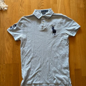 Ljusblå pikétröja från Polo Ralph Lauren - Säljer en ljusblå pikétröja från Polo Ralph Lauren med klassisk krage, knappar och stor broderad logga på bröstet. Tröjan har korta ärmar och en broderad siffra på ena ärmen. Modellen är custom slim fit.