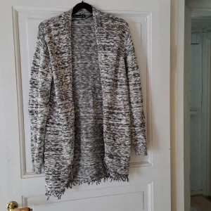 Gråmelerad stickad kofta - Säljer en lång, öppen gråmelerad stickad kofta med lång ärm. Koftan har ett avslappnat fall och fransiga kanter nedtill. Perfekt att slänga på sig över en t-shirt eller topp.