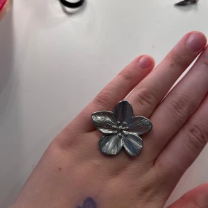 Stor silverfärgad blomring - Säljer en statementring i silverfärgat material med en stor blomformad design. Ringen har tydliga detaljerade blomblad och sitter snyggt på fingret. Perfekt för dig som vill sticka ut med dina accessoarer.