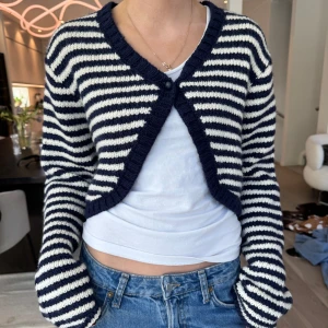 Randig croppad kofta i blått och vitt - Säljer en stickad, croppad kofta med marinblå och vita ränder. Koftan har lång ärm, v-ringning och en knapp framtill. Perfekt att slänga över en t-shirt för en avslappnad look. Storlek S och är knappt använd. I mycket bra skick! Säljs inte längre i butik🩷