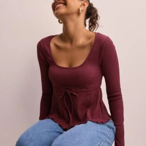Vinröd ribbad topp med knyt - Säljer en vinröd långärmad topp med ribbad struktur och knyt i midjan. Toppen har en djup rundad urringning och volangkant nertill. Perfekt för dig som gillar en feminin och trendig stil.