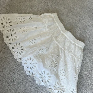 Vit kjol med broderade blommor och volangkant - Supersöt vit kjol med bred resår i midjan och broderade blommor över hela tyget. Kjolen har volangig nederkant med fina utskurna detaljer som ger en romantisk känsla. Perfekt för varma dagar!