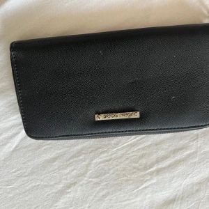 Svart plånbok från Björn Borg - Snygg svart plånbok från Björn Borg med stilren design och silverfärgad logga framtill. Plånboken har dragkedja runtom, flera kortfack och ett myntfack med dragkedja. Perfekt för att hålla ordning på dina kort och kontanter.