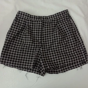 Svarta rutiga shorts - Svarta shorts från Monki med vitt rutmönster. De har råa kanter nertill och fickor både fram och bak. Klassisk passform med hällor för bälte och knappstängning framtill. Perfekta för en trendig och avslappnad look.