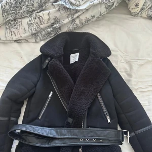 Svart mocka teddyjacka med bälte från Stradivarius - Säljer en svart teddyjacka från Stradivarius med stora slag, dragkedjor och bälte i midjan. Jackan har långärmade ärmar och är fodrad med mjuk teddy på insidan. Perfekt för kyliga dagar och ger en cool look. Använd 3 gånger, säljer eftersom att den är för stor på mig och aldrig kommer till användning :)