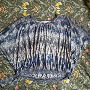 Blåvit mönstrad topp från Izabella - Säljer en luftig blåvit topp från Izabella med grafiskt mönster och knytning i sidan. Toppen har korta ärmar och rund halsringning, perfekt för en avslappnad stil.