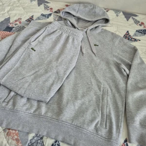 Lacoste dress - Säljer en ljusgrå dress från Lacoste med klassisk krokodillogga på bröstet. Tröjan har huva med dragsko och en ficka med dragkedja på sidan. Ett par byxor som machar med hoodien också. Mjuk och bekväm, perfekt för chill eller träning. Byxorna och hoodin är storlek L med det är slim fit så det sitter tight, rekommenderar den helst till storlek M 