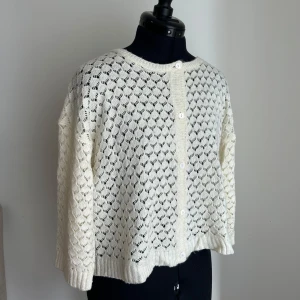 Vit stickad kofta med hålmönster - Säljer en söt vit stickad kofta med hålmönster och rund hals. Koftan har något 3/4 ärmar, ribbade muddar och knäpps med vita knappar framtill.