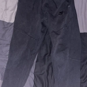 Svarta mjukisbyxor från Nike - Svarta mjukisbyxor från Nike med diskret logga på vänster lår. Byxorna har dragsko i midjan och en bakficka med dragkedja. Perfekta för en avslappnad stil eller träning.