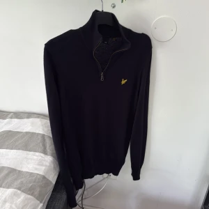 Mörkblå halvzip tröja från Lyle & Scott - Mörkblå långärmad tröja från Lyle & Scott med halv dragkedja och gul broderad logga på bröstet. Tröjan har ribbade muddar och är i ett mjukt material som passar perfekt till vardags.