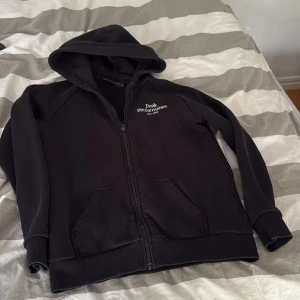 Svart hoodie från Peak Performance - Svart hoodie med dragkedja från Peak Performance. Tröjan har huva, två fickor framtill och vit broderad logga på bröstet. Perfekt för en avslappnad stil.